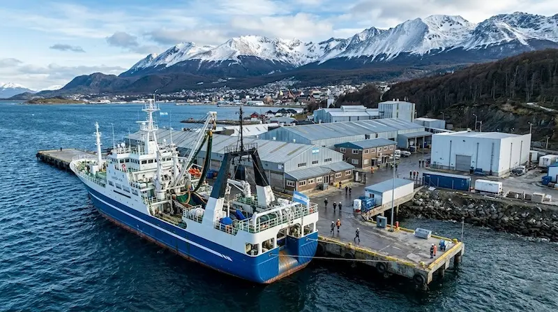 Barco pesquero amarrado en un muelle industrial de Ushuaia, con instalaciones portuarias y montañas nevadas al fondo.