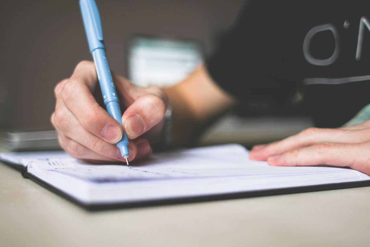 Persona sosteniendo bolígrafo azul escribiendo en el cuaderno.