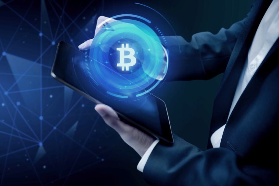 Persona interactuando con un holograma azul del símbolo de Bitcoin en una tablet, representando el control digital de las criptomonedas.
