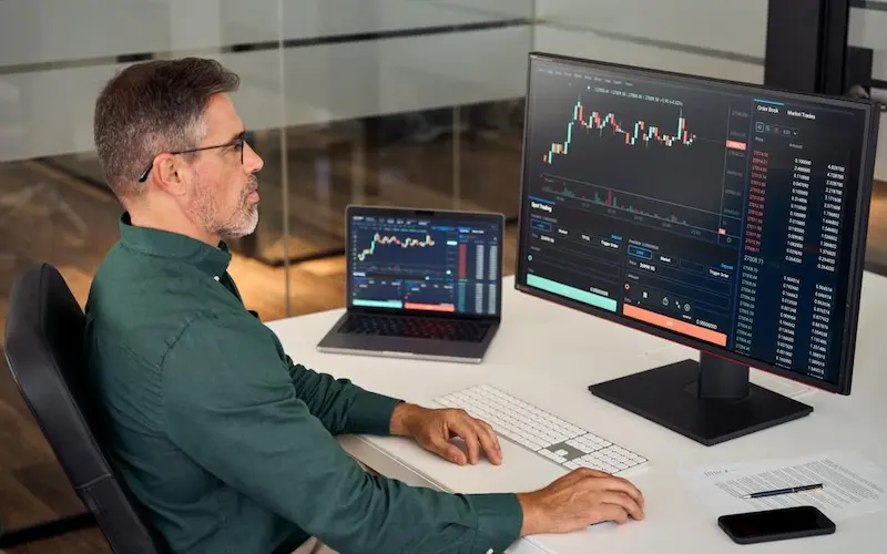 Hombre operando criptomonedas con trading bot en computadora y laptop