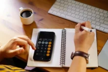 Persona calculando finanzas en un escritorio con calculadora del teléfono y cuaderno, concepto de tasa de interés real y costo del préstamo.