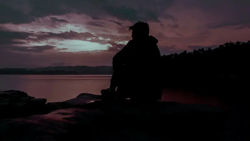 Silueta de un viajero sentado junto a un lago al atardecer