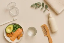 Naturaleza muerta horizontal con alineador dental en estuche, cepillo, peine de madera, serum capilar, vaso de agua y plato con salmón, palta y nueces sobre fondo beige.