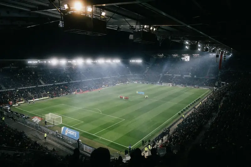 Vista aérea de un estadio de fútbol iluminado durante un partido con equipos formados en el centro del campo
