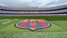 Primer plano a ras de suelo del escudo del FC Barcelona pintado en el césped del estadio Camp Nou vacío bajo un cielo nublado.