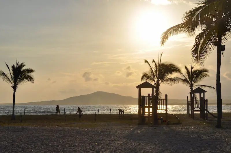 Atardecer en una playa tropical con palmeras y siluetas de personas frente al mar, con luz dorada y ambiente tranquilo
