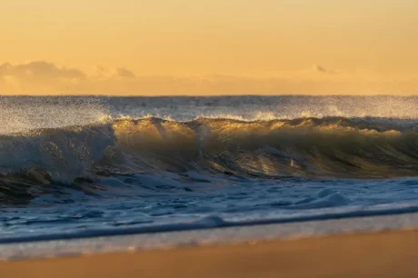 Ola rompiendo al atardecer en la costa, con espuma y luz dorada sobre el mar