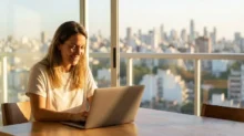 Persona trabajando con una notebook desde casa, con vista urbana, en un contexto de trabajo remoto y economía digital.