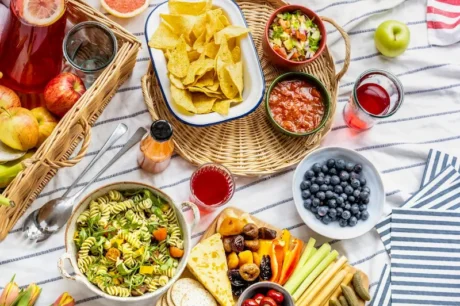 Picnic con ensalada de pasta, nachos con salsas, frutas frescas y bebidas sobre una manta a rayas.