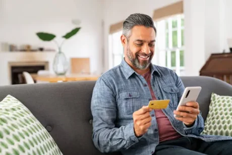 Hombre sentado en un sofá mira su celular y sostiene una tarjeta de crédito.