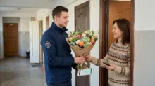Courier hands a bouquet to a woman at an apartment door while she holds a phone.