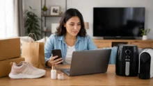 Mujer compara compras online con notebook y celular junto a zapatillas, cajas y electrodomésticos.