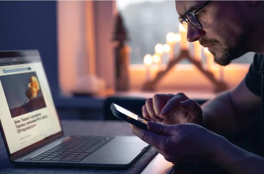 Hombre usando un celular junto a una notebook con contenido digital en pantalla