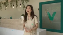 Profesional sanitaria en una clínica con recepción, plantas colgantes y logo verde en una puerta de vidrio