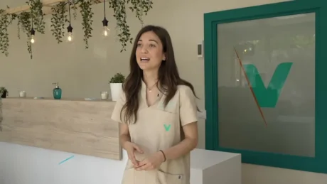 Profesional sanitaria en una clínica con recepción, plantas colgantes y logo verde en una puerta de vidrio