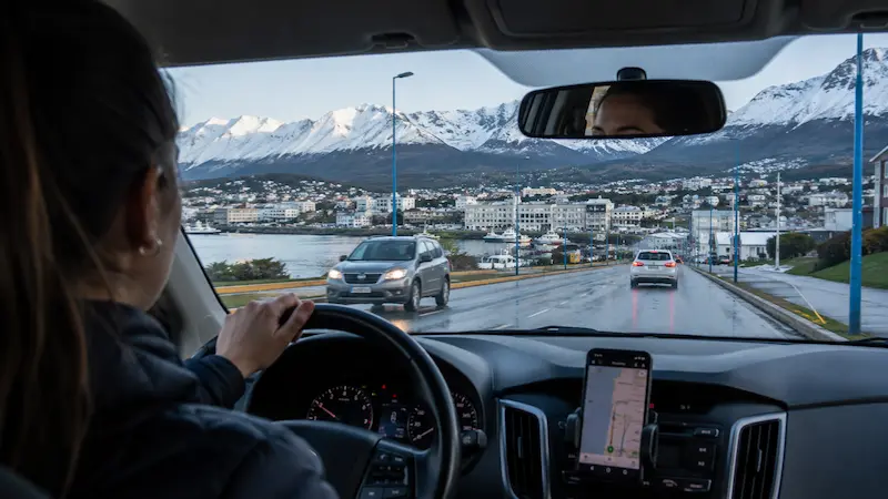 Conductora maneja por una ciudad costera con montañas nevadas de fondo