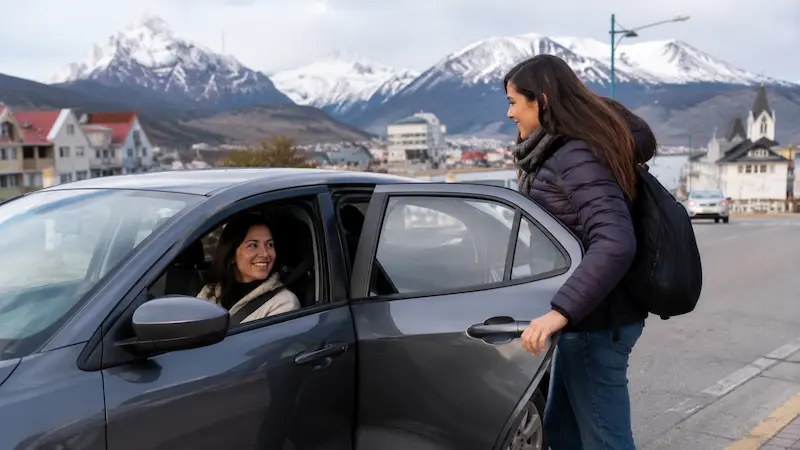 Mujer sube a un auto con conductora en una calle con montañas nevadas de fondo