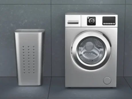 Lavarropas automático de carga frontal junto a un cesto de ropa en un lavadero gris.