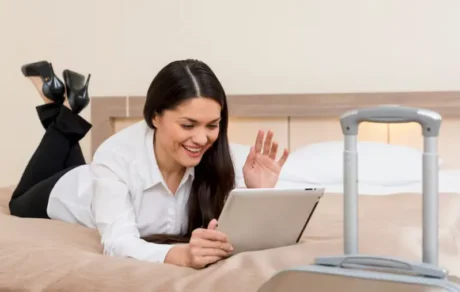 Mujer sonriente acostada en una cama consulta una tablet junto a una valija