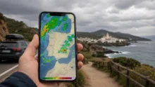 Teléfono con un radar de lluvia sobre España frente a una ruta y una localidad costera bajo cielo nublado