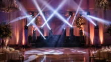 Salón de eventos con pista de baile, cabezales móviles y haces de luz sobre un escenario