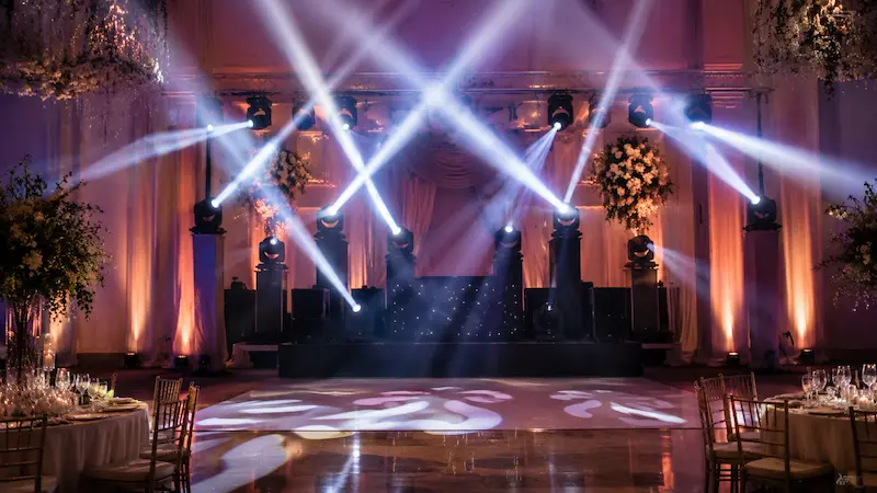 Salón de eventos con pista de baile, cabezales móviles y haces de luz sobre un escenario